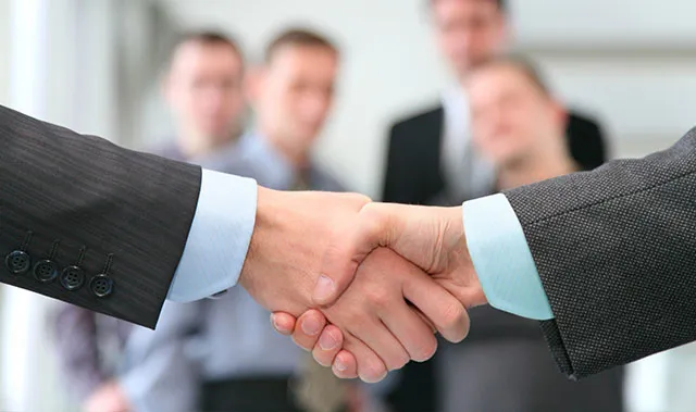 Dois empresários apertando as mãos em frente a um grupo de pessoas.